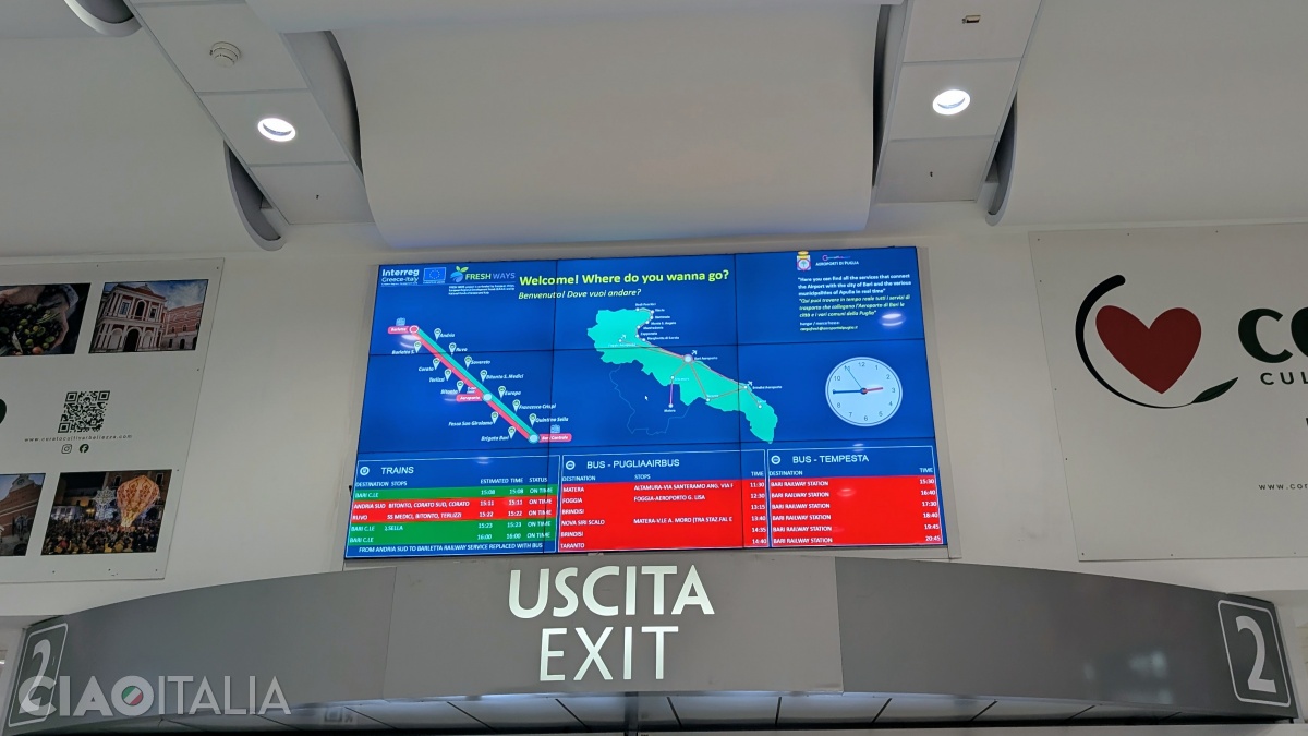 The bus and train schedule is displayed on a digital screen above the airport exit doors.